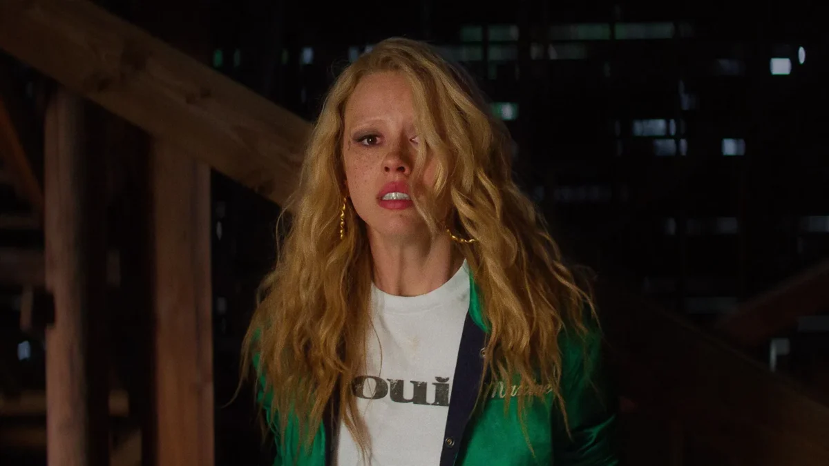 Mia Goth as Maxine Minx with long wavy blond hair, a cut on her cheek and a green jacket, in a still from MaXXXine