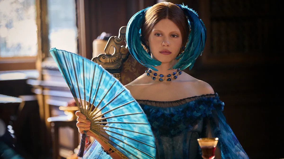 Mia Goth as Elizabeth in a blue gown and blue feathered headpiece, holding a blue fan, in a still from Frankenstein