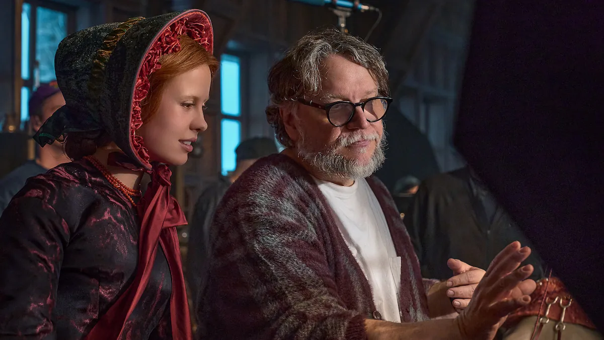 Mia Goth in a red-trimmed bonnet and period dress listening to Guillermo del Toro, who wears glasses and a cardigan, on the Frankenstein set