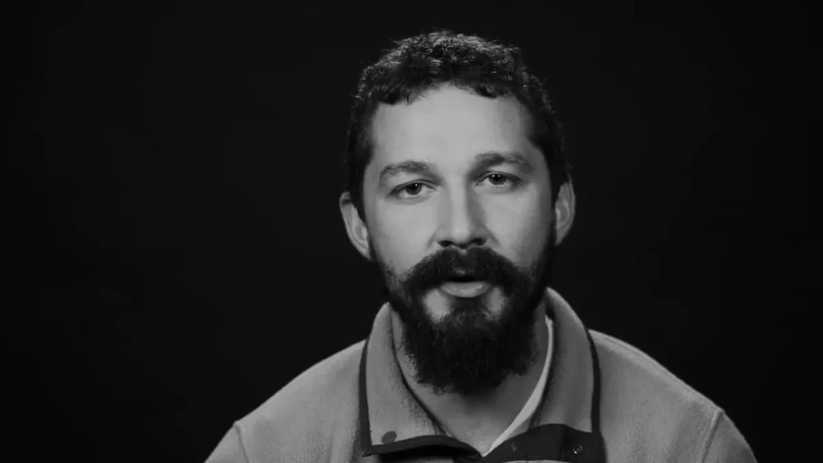 Shia LaBeouf in black and white, with a beard and a fleece pullover, speaking to the camera