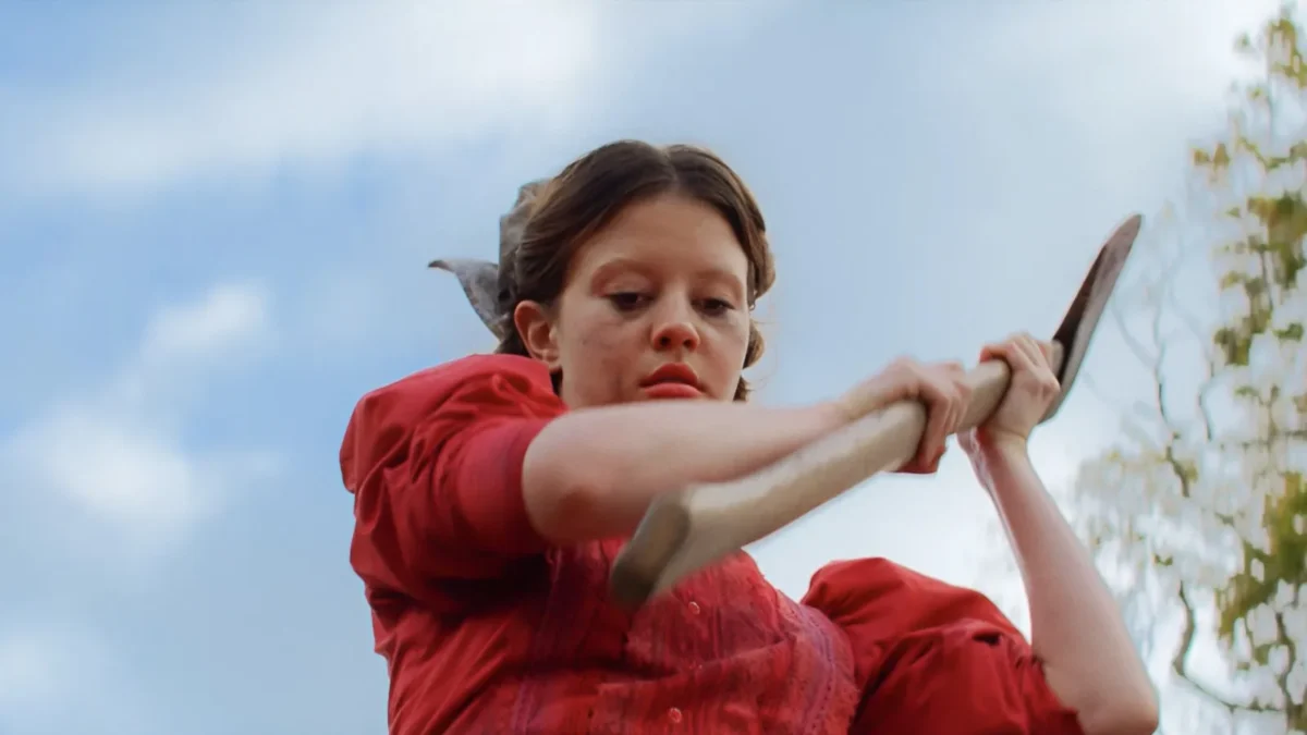 Mia Goth as Pearl in a red dress swinging an axe against a blue sky, in a still from Pearl