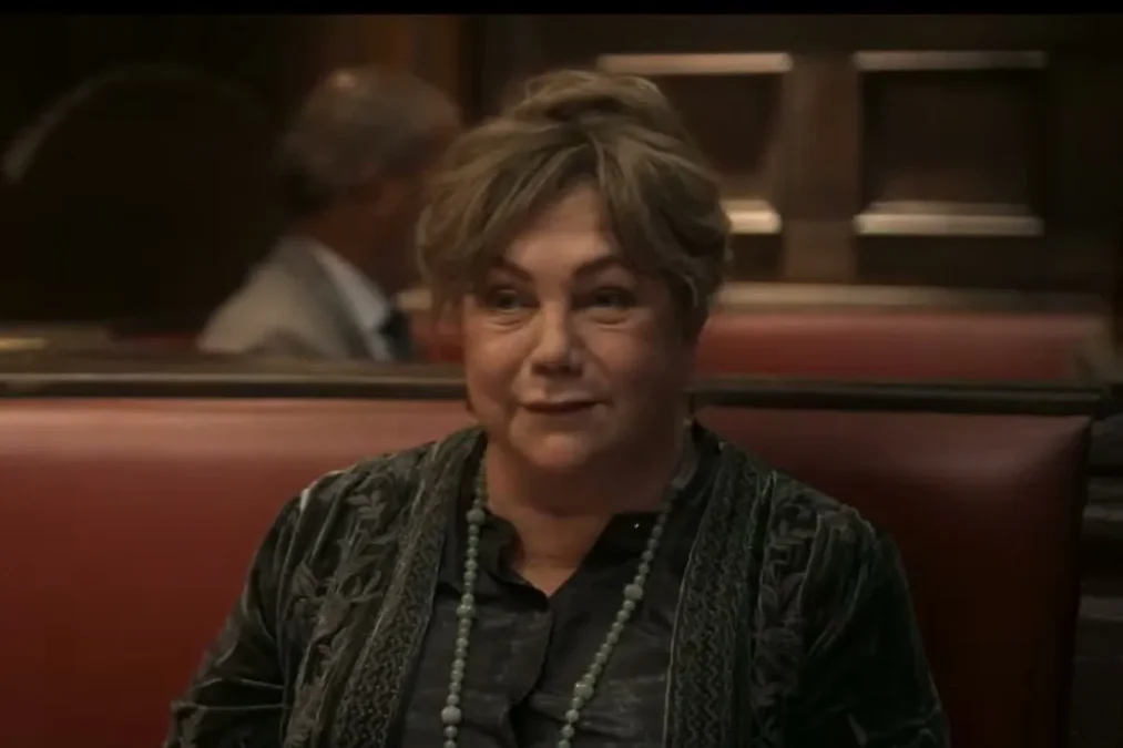 Kathleen Turner in a dark green jacket and long beaded necklace sitting in a red restaurant booth in The Kominsky Method