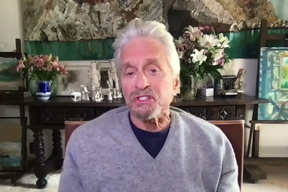 Michael Douglas in a gray sweater speaking on a video call in front of paintings and flowers