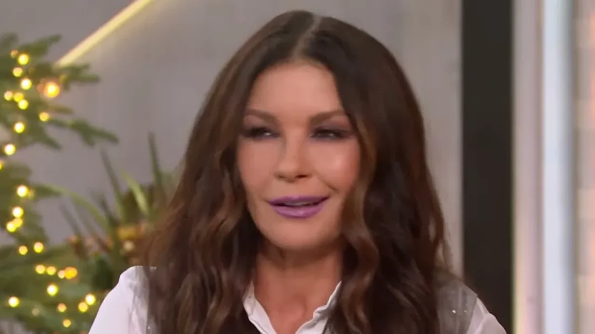 Catherine Zeta-Jones with long wavy dark hair in a white blouse on a talk show couch