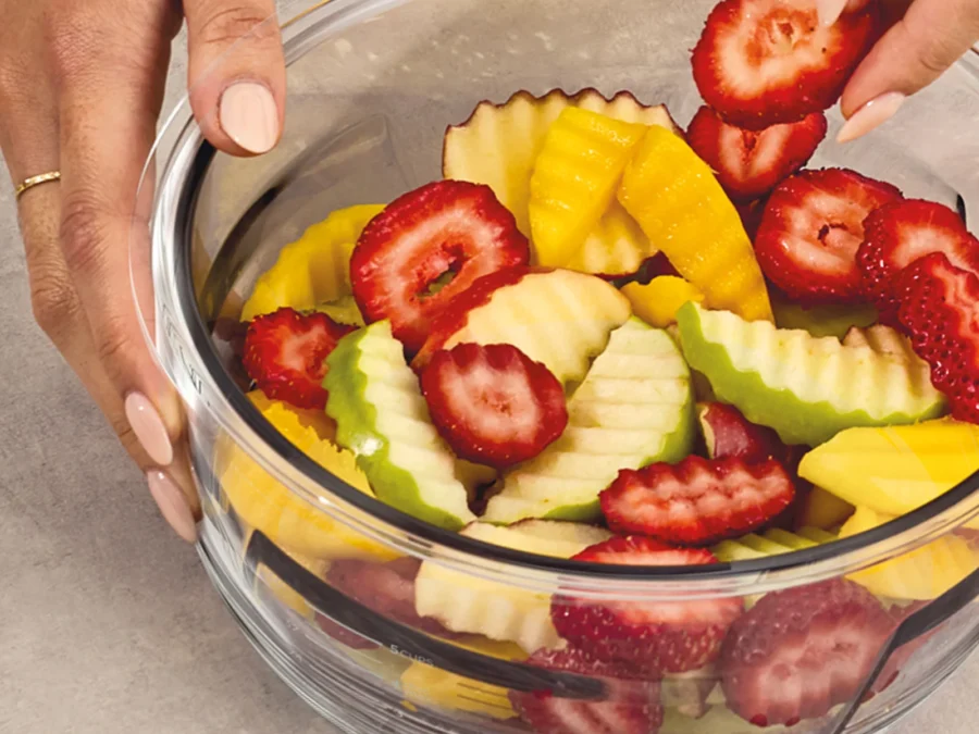 Hands holding a Ninja PREPi glass bowl full of crinkle-cut apples, mango and sliced strawberries