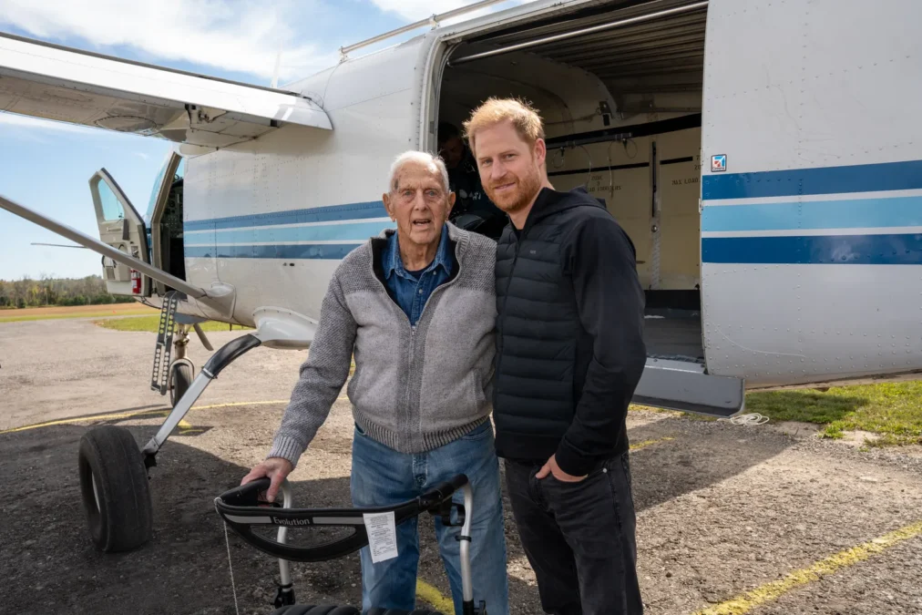 Prince Harry in a dark puffer vest standing next to World War II veteran Ed Marshall beside a small plane on an airfield