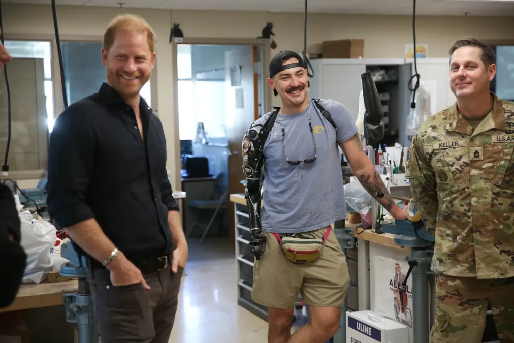 Prince Harry in a black shirt smiling next to two service members in a prosthetics workshop