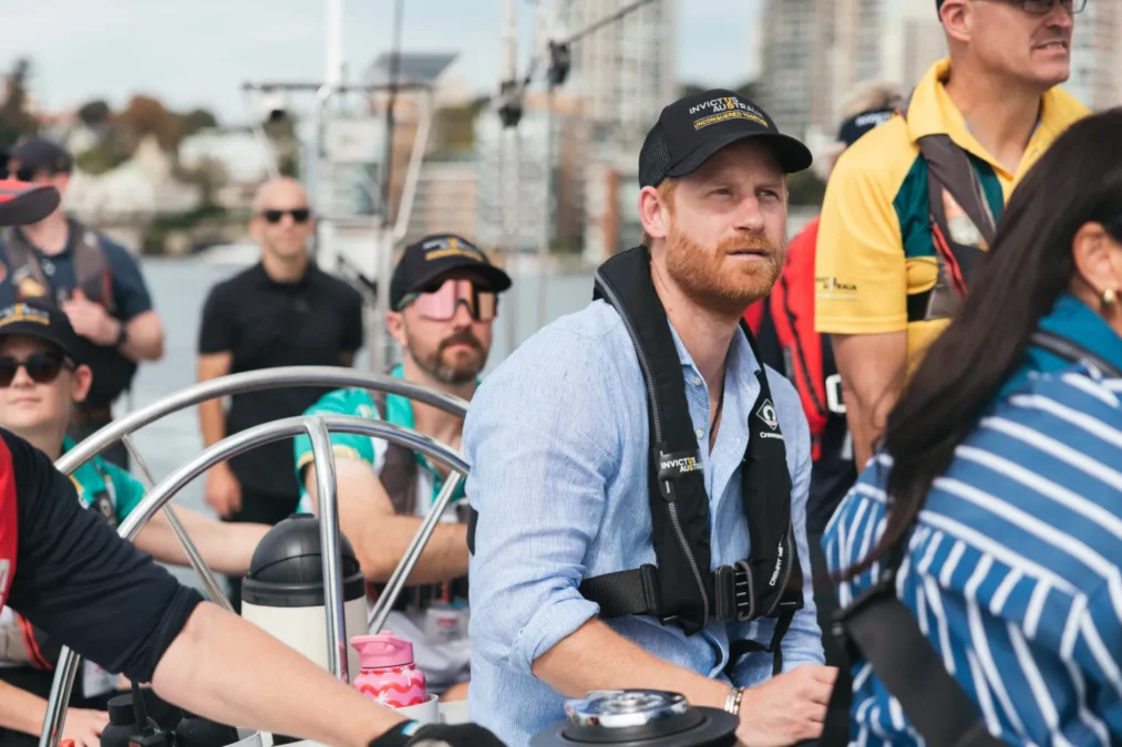 Prince Harry in a cap and life vest smiling on board a sailboat with other sailors