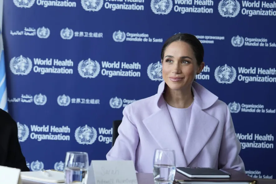 Meghan in a lilac blazer seated at a table in front of a World Health Organization backdrop