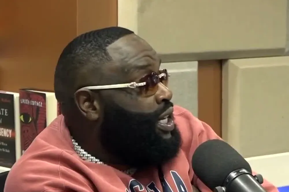 Rick Ross in a pink sweatshirt and tinted glasses speaking into a microphone at The Breakfast Club