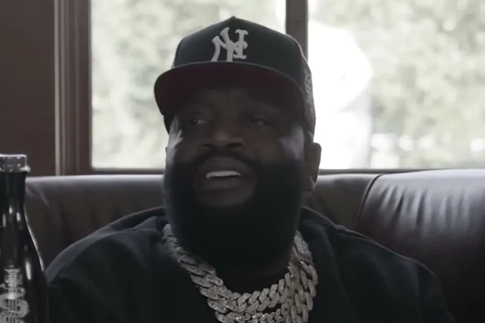 Rick Ross in a New York cap and a thick silver chain speaking at a desk in a wood-paneled office