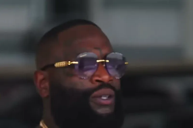 Rick Ross in a black T-shirt and gold chain with tinted glasses smiling in an airplane hangar