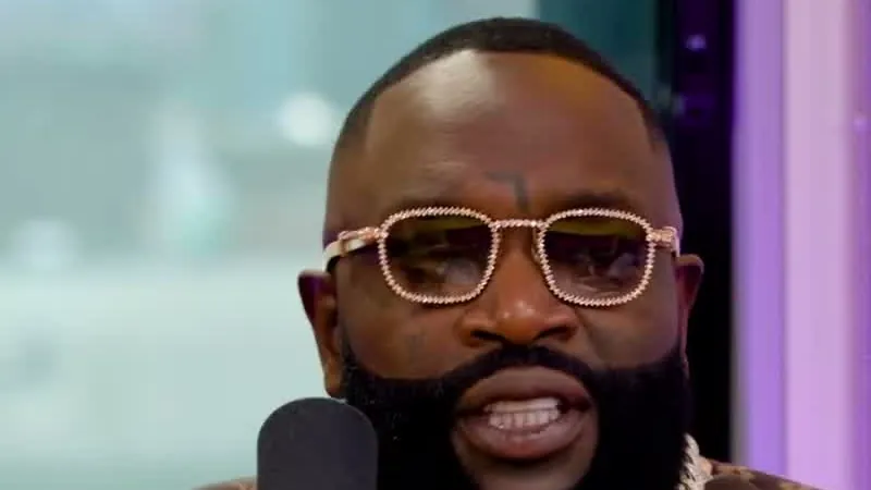 Rick Ross in pink-framed glasses and a brown Louis Vuitton-print shirt speaking into a microphone in a radio studio