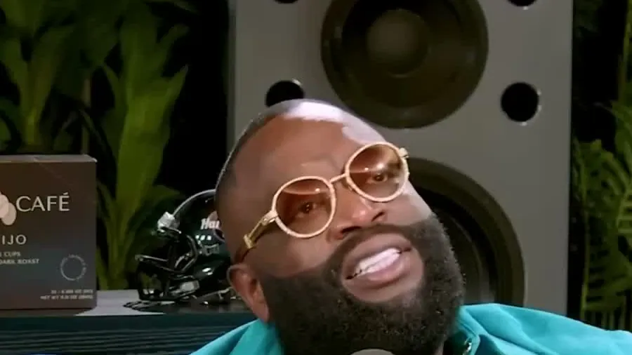 Rick Ross in a turquoise jacket and gold-framed glasses laughing on a blue couch