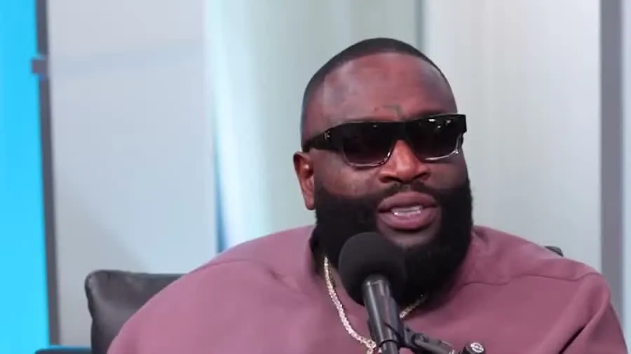 Rick Ross in dark sunglasses and a mauve shirt speaking into a microphone on SiriusXM's Hip Hop Nation set