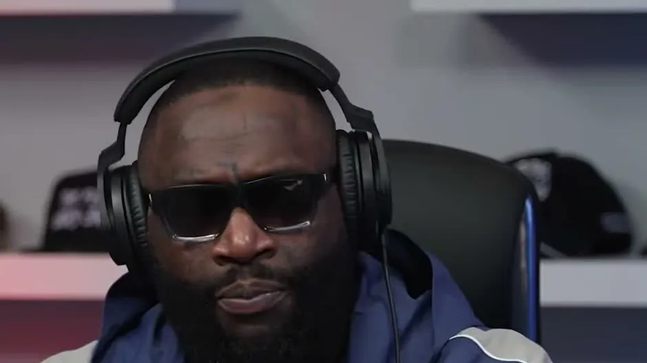 Rick Ross in dark glasses and headphones wearing a blue track jacket with a wall of caps behind him