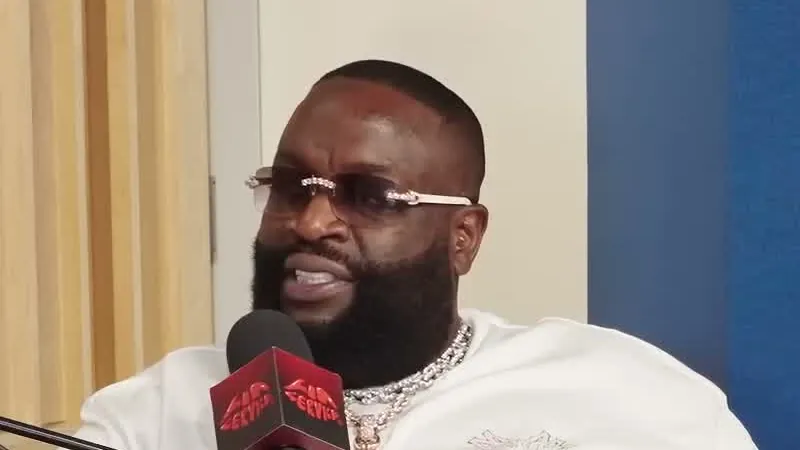 Rick Ross in a white T-shirt and tinted glasses sitting back on a leather couch holding a drink