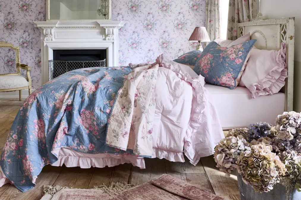 A blue and pink floral quilt layered over a pale pink quilt and ruffled pink sheets on a bed in a room with floral wallpaper and a white fireplace