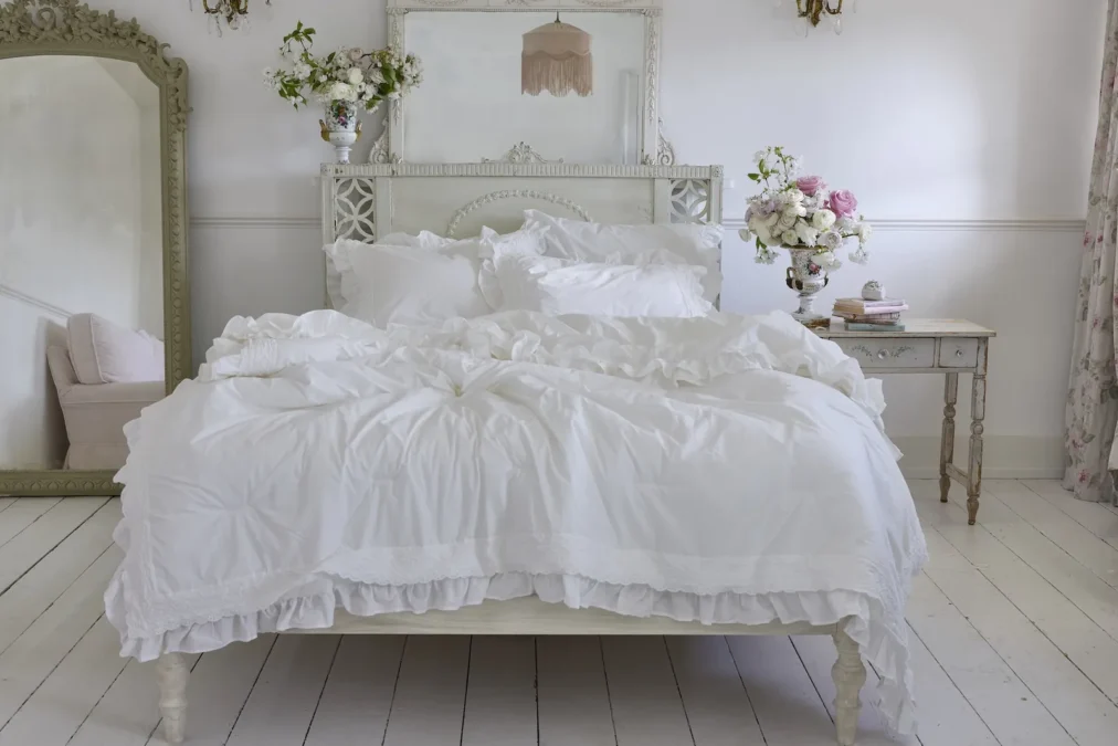 A white bed made with white ruffled bedding and pillows in a pale room with a vintage mirror and side table