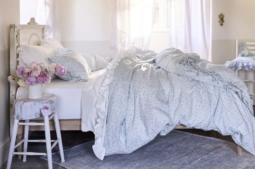 A blue and white small-print floral duvet with a ruffled edge on a bed beside a white stool holding pink roses