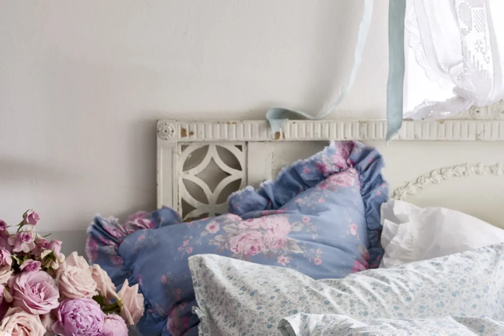 A blue floral pillow with a ruffled edge and pink roses printed on it, against a white carved headboard
