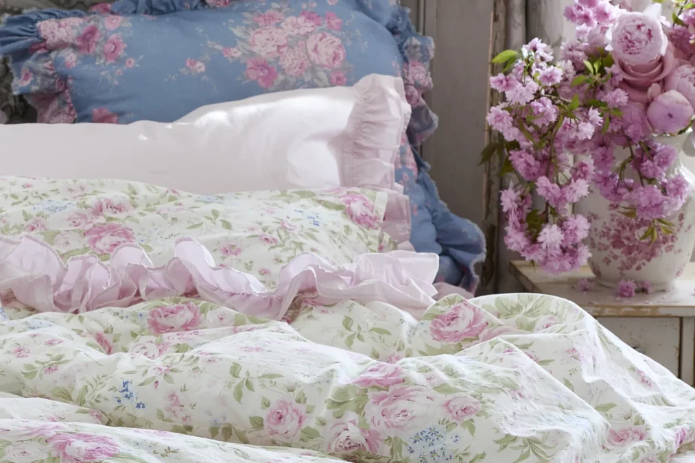 A pink and green rose print duvet with a pink ruffled pillowcase and a blue floral pillow, next to a vase of pink flowers