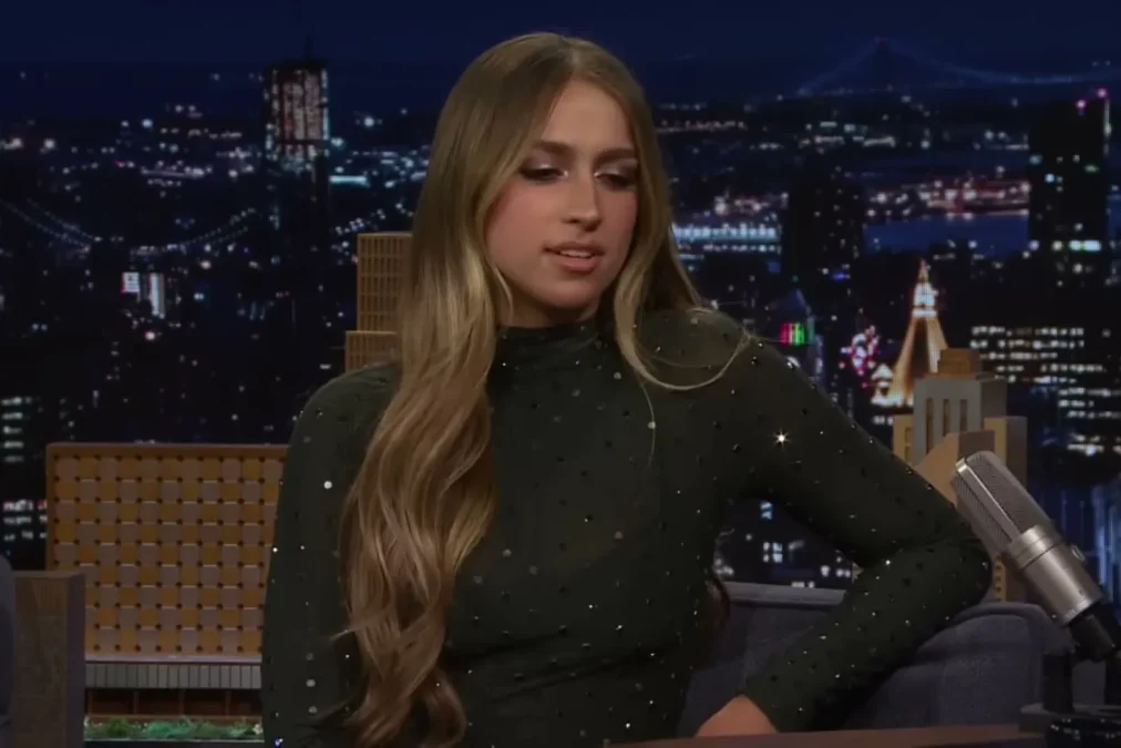 Tate McRae with long highlighted hair in a dark sparkly dress sitting on the couch on The Tonight Show set