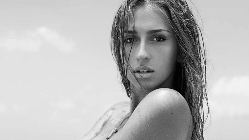 Black-and-white portrait of Tate McRae with wet hair looking at the camera, one hand at her neck, against a cloudy sky