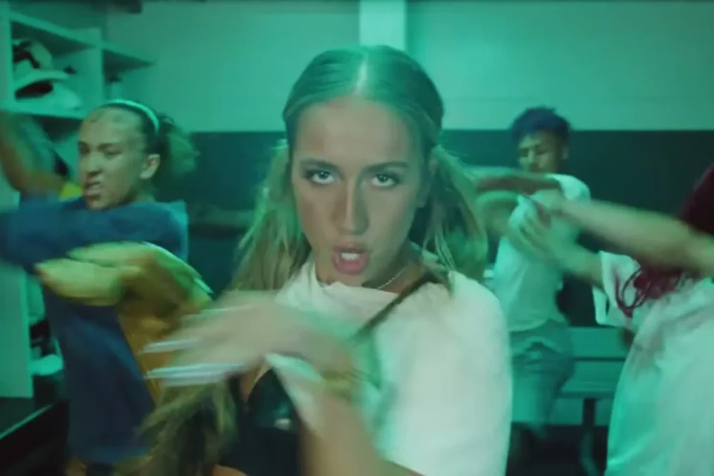 Tate McRae in a white T-shirt and a black top staring at the camera with dancers around her in a locker room