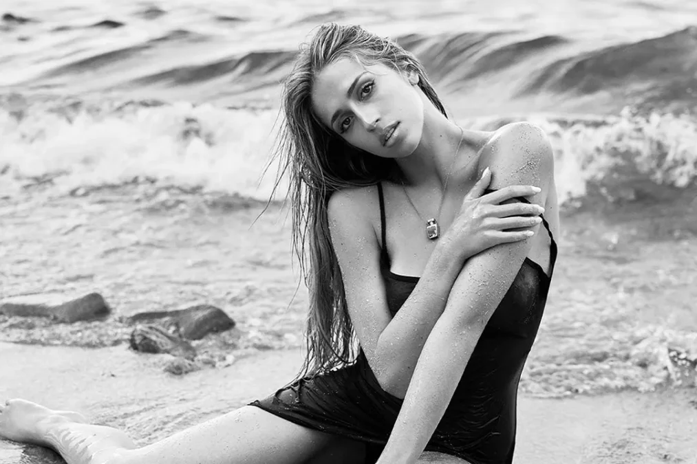 Black-and-white photo of Tate McRae sitting in the surf in a wet black slip dress looking at the camera, with waves behind her