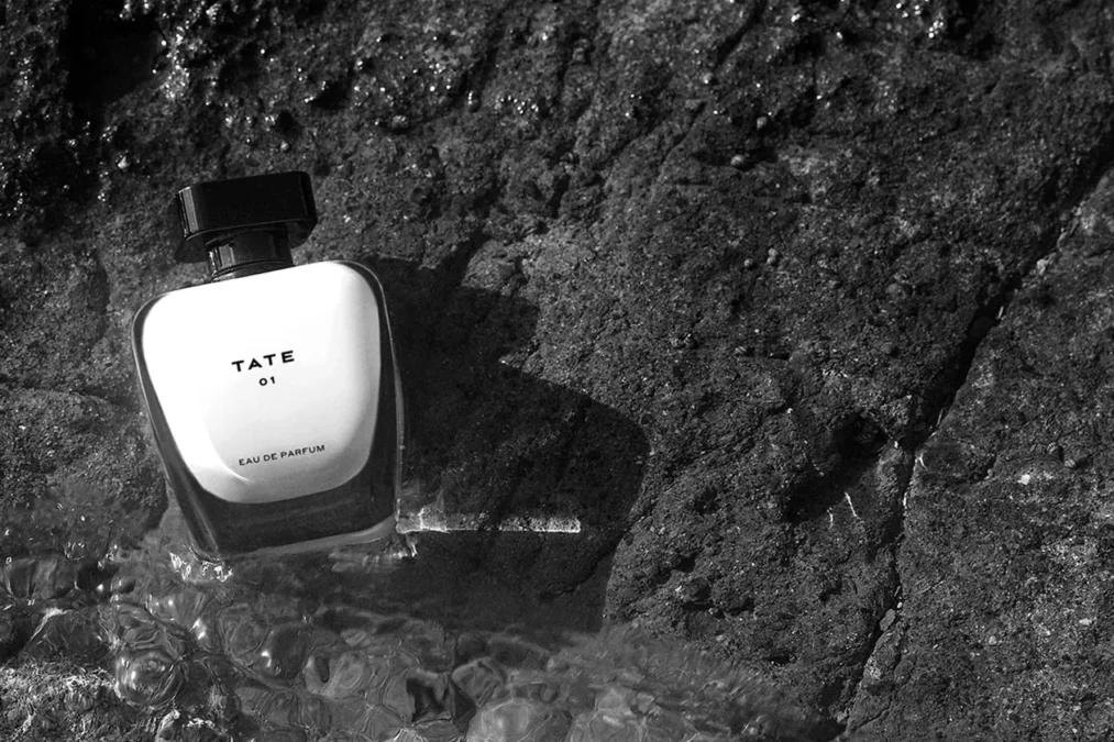 The TATE 01 eau de parfum bottle resting on dark volcanic rock with water around it, in black and white