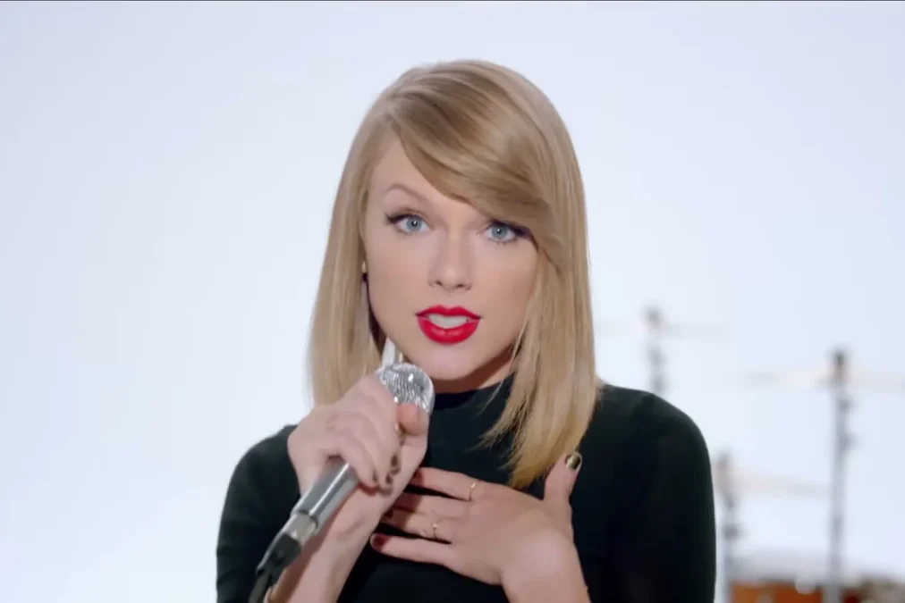 Taylor Swift with a short blond bob and red lipstick singing into a microphone in the Shake It Off music video