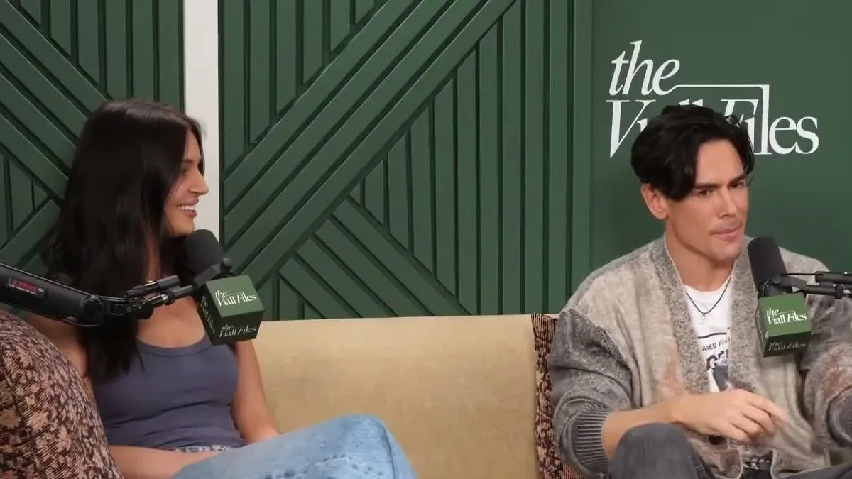 Victoria Lee Robinson, with long dark hair, smiling on a beige couch next to Tom Sandoval in a gray cardigan, on a podcast set