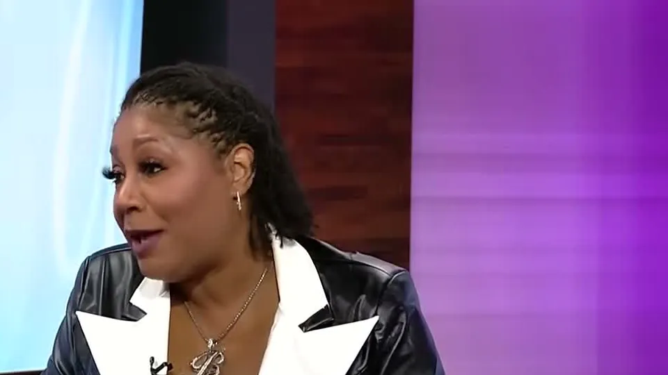Trina Braxton in a black leather jacket with white lapels and a script B necklace speaking on a TV set