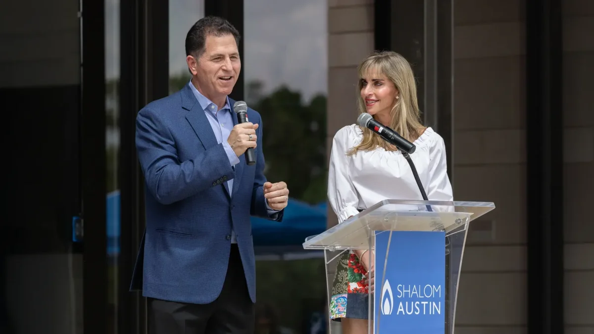 Michael Dell speaking into a handheld microphone next to Susan Dell, in a white blouse, at a clear lectern outdoors