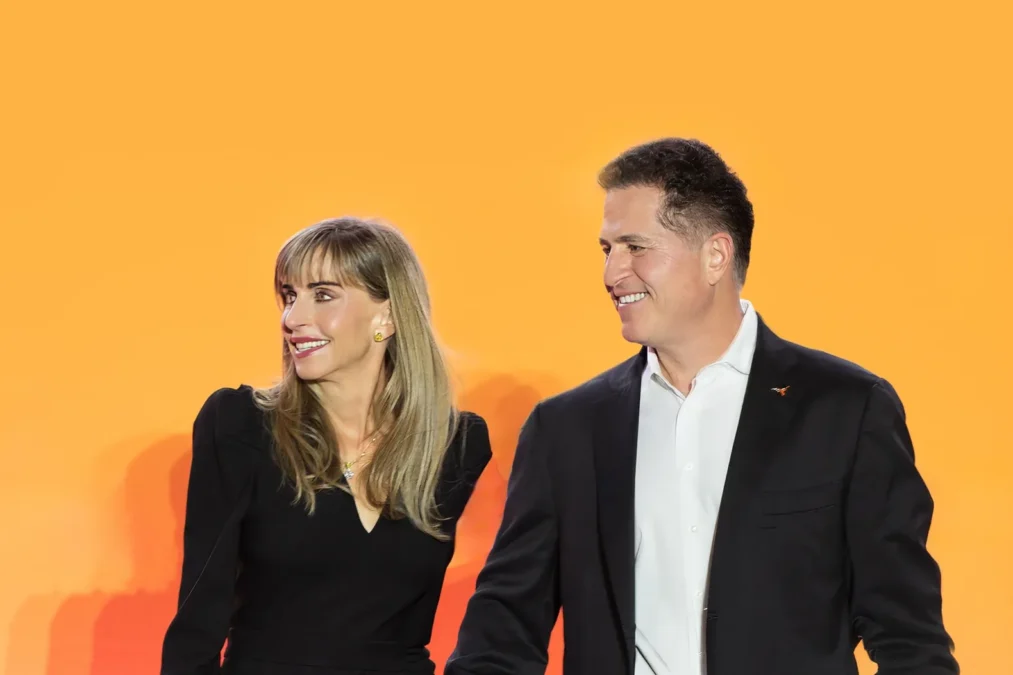 Susan Dell in a black dress smiling and looking at Michael Dell, in a dark blazer, against an orange background