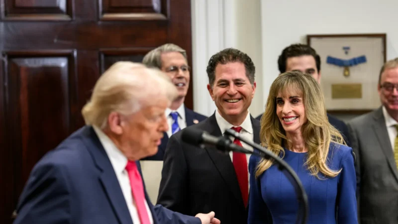 President Trump at a lectern in the Roosevelt Room, with Michael Dell and Susan Dell, in a blue dress, smiling behind him